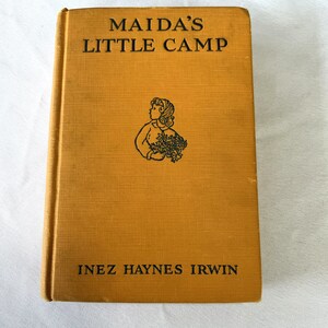 May include: A vintage book titled "Maida's Little Camp" by Inez Haynes Irwin. The cover is a yellow-brown color with a black and white illustration of a girl holding flowers.