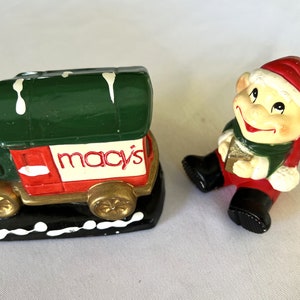 May include: A green and red toy delivery truck with the word "Macy's" on the side, and a smiling elf figurine wearing a red hat and green clothing.