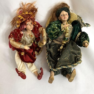 May include: Two decorative dolls with detailed costumes. One doll wears a red and gold outfit with a feathered headdress. The other doll is dressed in a green and gold ensemble with gold accents and a gold halo. Both dolls have painted faces.