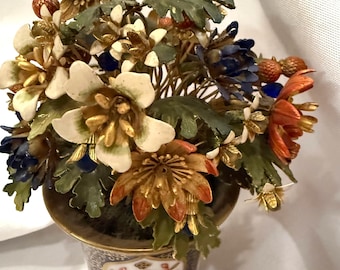 May include: A decorative floral arrangement in a small, ornate vase. The flowers are a mix of colours, including gold, white, blue, and orange. The vase has a gold rim and base, with a detailed floral pattern. The arrangement includes leaves and berries.