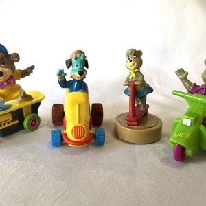 May include: Set of four plastic toy figures of bears and a dog riding on a skateboard, scooter, car, and a motor scooter. The figures are all wearing colorful clothing and have a cartoonish style.