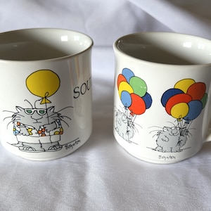May include: Two white ceramic mugs with cat illustrations. One mug features a cat holding a yellow balloon with the word "SO" on the mug. The other mug features a cat holding a bunch of colorful balloons. Both mugs have the word "Boynton" at the bottom.