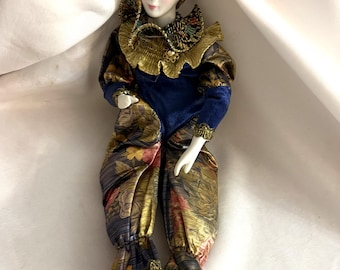 May include: A porcelain doll in a harlequin outfit. The doll features a blue velvet hat and bodice, accented with gold trim. The trousers display floral patterns in gold, red, and blue. The doll has a painted face with blue eyeshadow and black shoes.