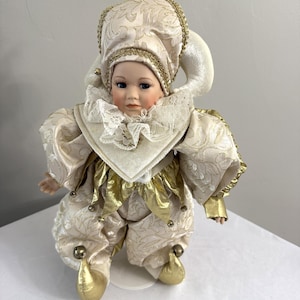 May include: A porcelain doll dressed as a jester, featuring a cream-colored outfit with gold accents. The doll has a detailed face with blue eyes and is wearing a hat with a bell. The jester's costume includes a ruffled collar and gold-colored bells on the shoes.