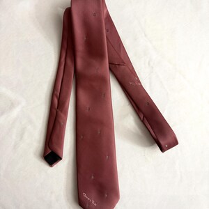 May include: A dusty rose-colored tie with a subtle pattern of small, square-shaped designs. The tie is made of a smooth, possibly silk, material and features the designer's signature near the tip. The tie is displayed on a white surface.