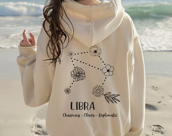 May include: A cream-colored hoodie with a black floral Libra zodiac constellation design on the back. The text "LIBRA Charming - Clever - Diplomatic" is printed below the design.
