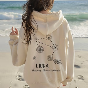 Puede incluir: Una sudadera con capucha de color crema con un dise&ntilde;o de constelaci&oacute;n zodiacal de Libra floral negro y el texto "LIBRA Charming - Clever - Diplomatic" en la espalda.