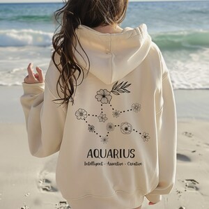 Puede incluir: Una sudadera con capucha de color crema con un dise&ntilde;o floral de la constelaci&oacute;n de Acuario y el texto "Aquarius, Intelligent - Assertive - Creative" en la espalda.