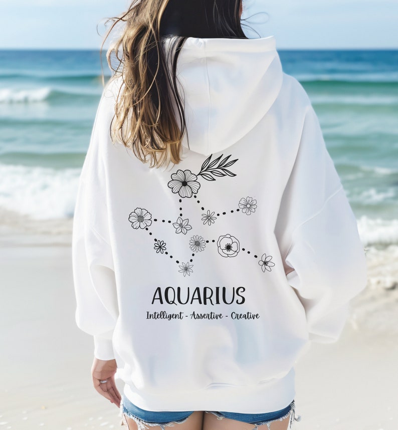 Puede incluir: Sudadera con capucha blanca con un dise&ntilde;o floral negro del signo zodiacal Acuario. El texto "Aquarius" y "Intelligent - Assertive - Creative" est&aacute; impreso debajo del dise&ntilde;o.