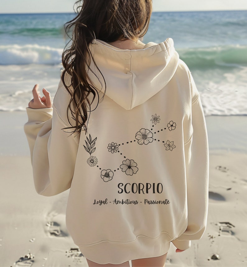 Puede incluir: Una sudadera con capucha de color crema con un signo zodiacal de Escorpio floral y el texto "Loyal - Ambicioso - Apasionado" impreso en la espalda.