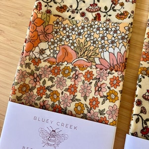 May include: A set of beeswax wraps featuring three distinct floral patterns in shades of orange, pink, and cream. The wraps are secured with a white paper band labelled BLUEY CREEK BEESWAX WRAPS with a bee illustration and the text NATURAL SUSTAINABLE HANDMADE.