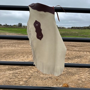 May include: A white leather chaps with brown leather trim and fringe. The chaps are hanging on a black metal fence.