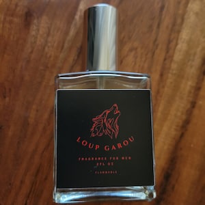 May include: A rectangular glass perfume bottle with a silver cap. The black label features a red wolf illustration and the words "LOUP GAROU FRAGRANCE FOR MEN 2FL OZ FLAMMABLE". The bottle is on a wooden surface.