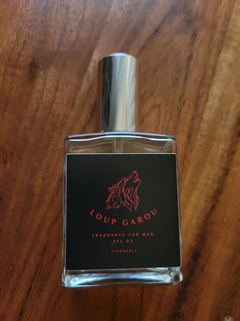 May include: A rectangular glass perfume bottle with a silver cap. The black label features a red wolf illustration and the words "LOUP GAROU FRAGRANCE FOR MEN 2FL OZ FLAMMABLE". The bottle is on a wooden surface.