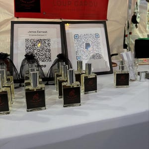 May include: A display of perfume bottles with black labels and silver caps. The bottles are arranged on a white table, with framed QR codes and a red banner that reads "LOUP GAROU". The brand name and a wolf logo are visible.