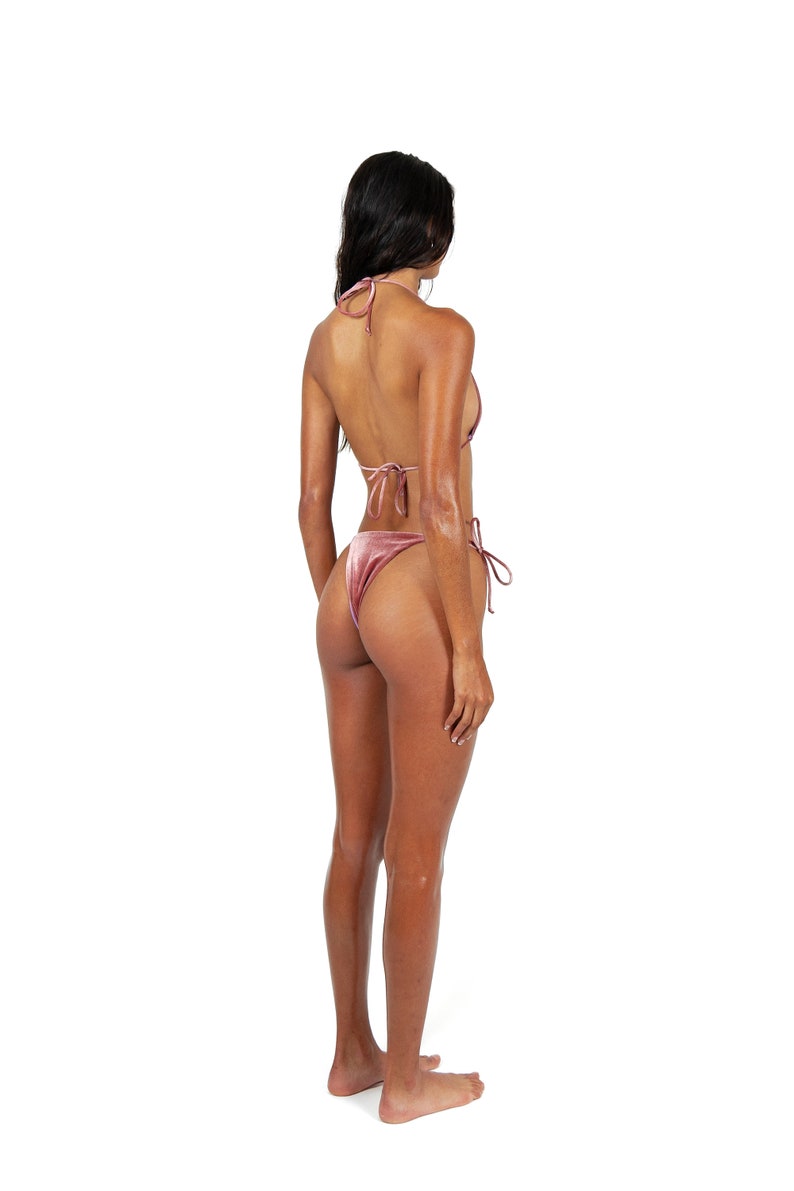 May include: A woman wearing a pink velvet bikini. The bikini top is a triangle style with thin straps. The bikini bottom is a high-cut style with ties on the sides.