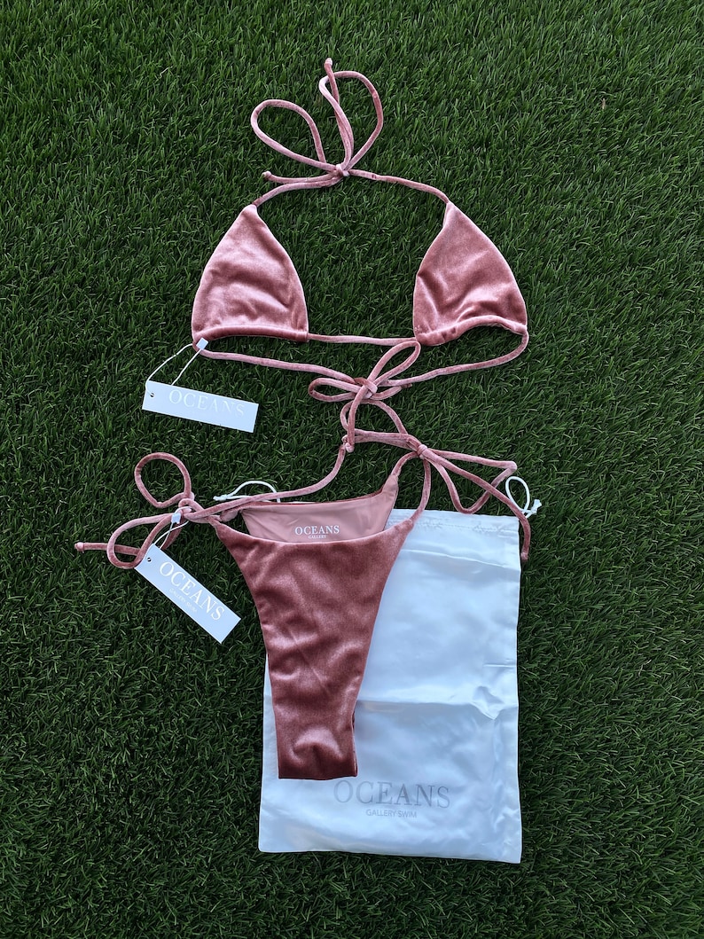 May include: A dusty rose velvet bikini set with a triangle top and a high-cut bottom. The bikini is laid out on a green grassy surface with a white drawstring bag and a white tag that says "OCEANS GALLERY SWIM".