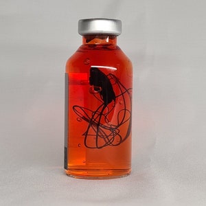 May include: A clear glass bottle with a silver cap, filled with a vibrant orange liquid. The bottle features a black abstract design. The bottle is likely for essential oils or a similar product.