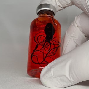 May include: A clear glass bottle filled with a vibrant red liquid. Inside, a black, abstract design is suspended. The bottle has a silver cap and is held by a gloved hand. The overall aesthetic is scientific or artistic.