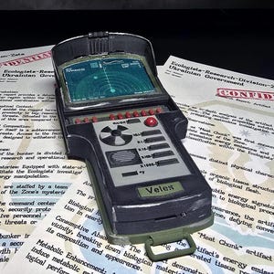 Op de afbeelding: Een zwart en olijfgroen elektronisch handapparaat met een scherm dat een radarinterface weergeeft, rode en zwarte knoppen en een label met de tekst Veles. Het apparaat rust op twee witte documenten met de markering Confidential.