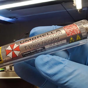 Op de afbeelding: Een zilveren T-Virus-injector van Umbrella Corporation, met het bedrijfslogo en de tekst "CAUTION". De injector bevat 50 ml vloeistof en heeft een rood-wit gevaarsymbool.