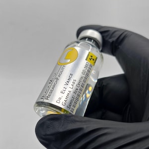 Peut inclure: Un flacon en verre transparent avec un bouchon argenté et une étiquette portant le texte "BLACK MESA RESEARCH FACILITY" et "SAMPLE: XEN CRYSTAL GG-3883". Le flacon est tenu par une main gantée.