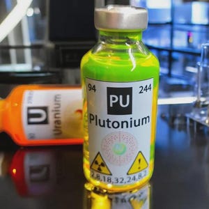 Può includere: Una fiala di vetro riempita con un liquido verde neon brillante con un'etichetta che riporta 94 PU Plutonium 244, un diagramma atomico circolare e due simboli triangolari gialli di pericolo radiazioni, accanto a una seconda fiala arancione Uranium.