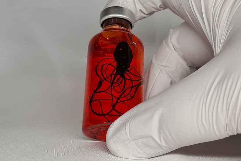 May include: A clear glass bottle filled with a vibrant red liquid. Inside, a black, abstract design is suspended. The bottle has a silver cap and is held by a gloved hand. The overall aesthetic is scientific or artistic.