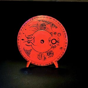 Peut inclure: Un disque rouge circulaire orné de motifs graphiques et de symboles noirs complexes sur toute sa surface. Le disque est fixé sur un petit support rouge, présentant un trou central et une série de motifs détaillés rappelant des marques stylisées.