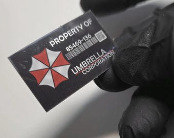May include: A rectangular black sticker featuring the white text PROPERTY OF, the number 85469-136, a barcode, a QR code, and the Umbrella Corporation logo, which consists of a red and white segmented umbrella icon with the text UMBRELLA CORPORATION.