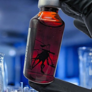 May include: A vial with a silver cap and a label with the text "SCP" is held by a gloved hand. The vial contains a dark red liquid and a black, abstract shape. The background is a blurred laboratory setting.