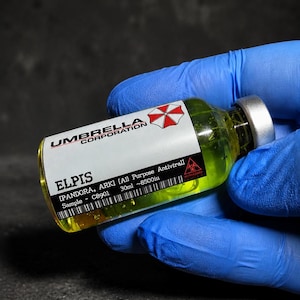 Peut inclure: Un flacon en verre transparent rempli de liquide jaune, avec un bouchon argenté et une étiquette blanche. L'étiquette porte le texte UMBRELLA CORPORATION, ELPIS, PANDORA, ARK, All Purpose Antiviral, Sample C8901, 30ml, 8500iu et un symbole biologique.