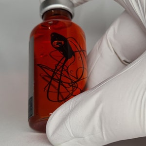 May include: A clear glass bottle filled with a reddish-orange liquid. The bottle has a silver cap and a black abstract design on the front. The bottle is held by a person wearing white gloves.