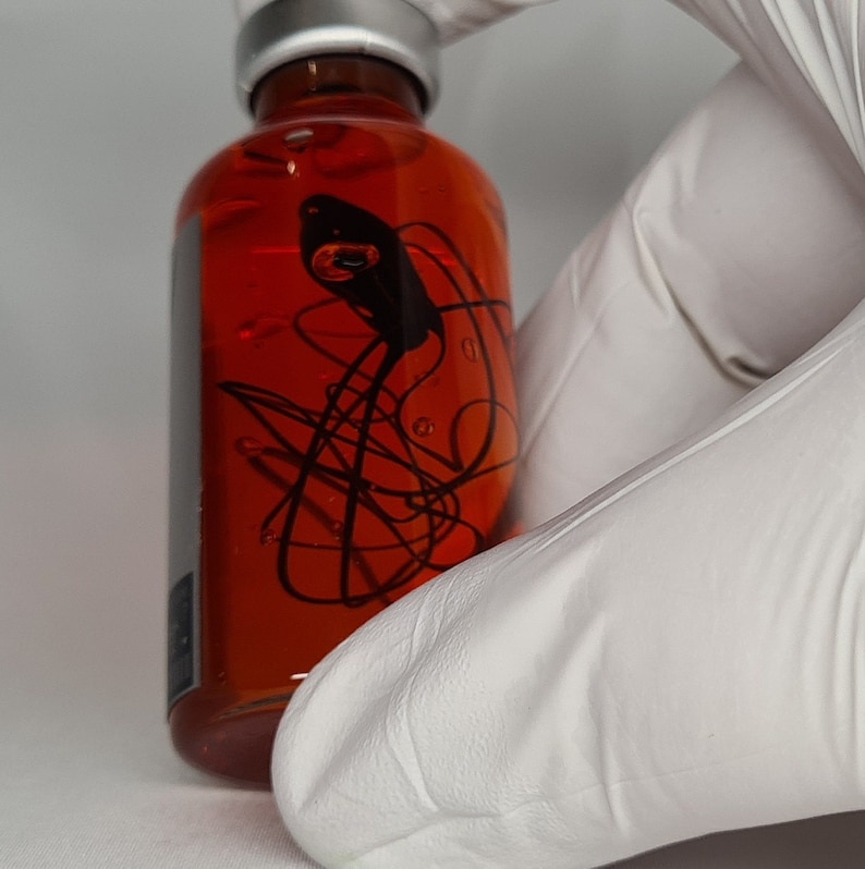 May include: A clear glass bottle filled with a reddish-orange liquid. The bottle has a silver cap and a black abstract design on the front. The bottle is held by a person wearing white gloves.