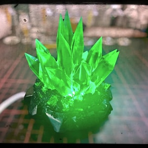 Może przedstawiać: Grupa jasnozielonych kryształów o ostrych, spiczastych krawędziach, wyrastających z ciemnej, teksturowanej podstawy. Kryształy są podświetlone, tworząc efekt świecenia. Obraz ma wygląd vintage, lekko wyblakły.