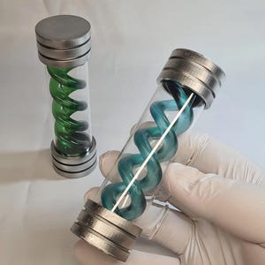 Op de afbeelding: Twee cilindrische glazen flesjes met metallic zilveren doppen en voetstukken. Eén flesje bevat een levendige groene spiraalstructuur, terwijl het andere flesje op de voorgrond een blauwgroene spiraalstructuur in de glazen buis heeft.