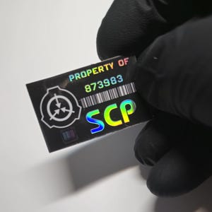 Könnte beinhalten: Ein rechteckiger holografischer Aufkleber mit dem Text "PROPERTY OF" über der Nummer "873983" und den Buchstaben "SCP" in einem Farbverlauf von Blau nach Gelb. Der Aufkleber hat auch ein Symbol und einen Barcode.