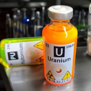 Może przedstawiać: Dwa szklane fiolki zawierające pomarańczowe i zielone płyny, oznaczone jako "Uran" i "Pluton". Pomarańczowa fiolka ma srebrną nakrętkę i białą etykietę z symbolem pierwiastka "U". Zielona fiolka leży. Obecne są symbole ostrzegawcze.
