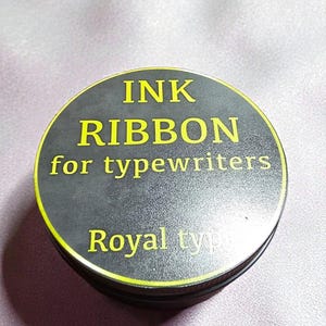 Könnte beinhalten: Runde, schwarze Tintenbanddose mit gelber Schrift. Der Text lautet "INK RIBBON for typewriters" und "Royal type". Die Dose hat einen gelben Rand und liegt auf einem hellvioletten Stoff.