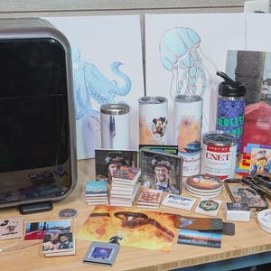 May include: A black eufy Make 3D printer sits beside a collection of printed items including cylindrical tumblers, square coasters, rectangular photo prints, a water bottle with CNET branding, and canvas art featuring an octopus and jellyfish.