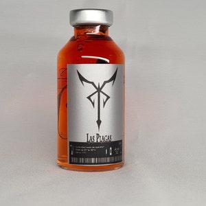 May include: A small glass bottle with a silver cap, filled with a bright orange liquid. A silver label displays a black emblem and the words "Las Plagas". The bottle has a barcode and other text.