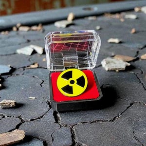 Pode incluir: Um alfinete com um símbolo de radiação amarelo e preto, exibido em uma caixa de plástico transparente com uma base vermelha. O alfinete está sobre uma superfície cinza escura rachada com detritos espalhados.