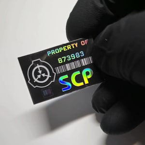 Könnte beinhalten: Ein rechteckiger schwarzer Aufkleber mit holografischem Finish, der den Text PROPERTY OF 873983, einen Strichcode und die Buchstaben SCP in großer, bunter Schrift zeigt. Links befindet sich ein kreisförmiges Logo mit drei Pfeilen zur Mitte.