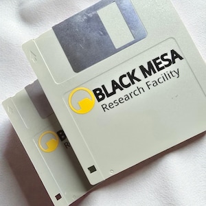Könnte beinhalten: Zwei hellgraue 3,5-Zoll-Disketten mit dem Text "BLACK MESA Research Facility" und einem gelben Logo. Die Disketten sind auf einer weißen Oberfläche gestapelt.