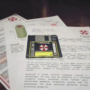 Puede incluir: Un disquete negro de 3,5 pulgadas con una etiqueta que muestra el logotipo y el texto de Umbrella Corporation, colocado sobre dos documentos impresos en blanco. Los documentos contienen texto técnico, diagramas y el logotipo rojo y blanco de Umbrella.