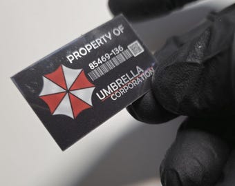 May include: A rectangular black and red tag with the text "PROPERTY OF" and "UMBRELLA CORPORATION". The tag features a barcode and the number "85469-136". The Umbrella Corporation logo is a red and white umbrella.