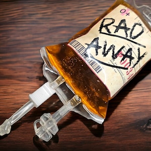Könnte beinhalten: Ein klarer Plastikbeutel gefüllt mit einer gelben Flüssigkeit, beschriftet mit "RAD AWAY" in schwarzer Schrift im Marker-Stil. Der Beutel hat angebrachte transparente Schläuche und Anschlüsse und liegt auf einer dunklen Holzoberfläche. Der Beutel hat auch einen Barcode und anderen Text.