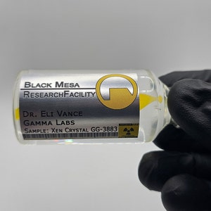 May include: A transparent glass vial containing yellow liquid, labelled "BLACK MESA RESEARCH FACILITY." The label also states "Dr. Eli Vance, GAMMA LABS, SAMPLE: Xen Crystal GG-3883." The vial is held by a gloved hand.