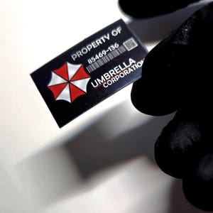 Puede incluir: Una tarjeta en blanco y negro con el texto "PROPERTY OF" y "UMBRELLA CORPORATION". La tarjeta presenta un logotipo de paraguas rojo y blanco y un código de barras. La tarjeta está sujeta por una persona que lleva un guante negro.