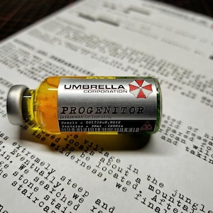 Op de afbeelding: Een klein glazen flesje gevuld met gele vloeistof, voorzien van een zilverkleurig etiket met de tekst UMBRELLA CORPORATION, PROGENITOR, STAIRWAY OF THE SUN, en bevat 30ml. Het flesje heeft een zilveren dop en een biohazard-symbool.
